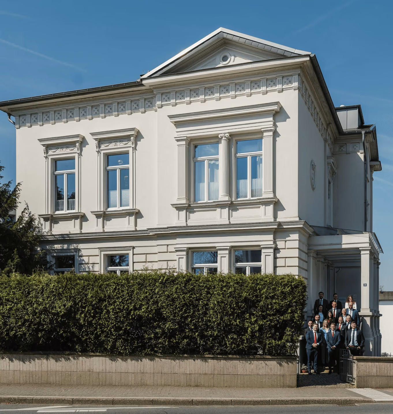 Gruppenfoto von zwanzig formell gekleideten Personen, die auf der Treppe eines historischen weißen Gebäudes mit grüner Hecke stehen.